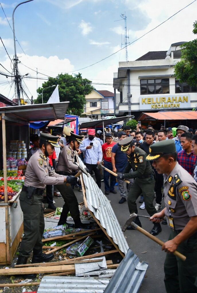 Petugas pemerintah daerah menggunakan palu dan alat berat membongkar warung kecil di pinggir jalan, warga sekitar berdiri menyaksikan dengan ekspresi tegang dan khawatir, latar belakang kantor kelurahan terlihat jelas, suasana dramatis namun terorganisir, gaya foto jurnalistik resolusi tinggi